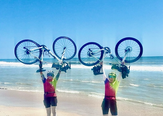 Two women by the ocean holding their bikes over their heads.