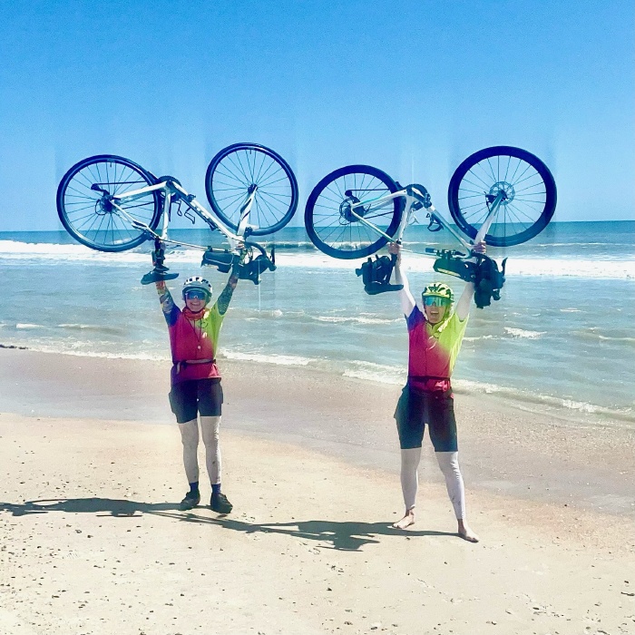 Two women by the ocean holding their bikes over their heads.