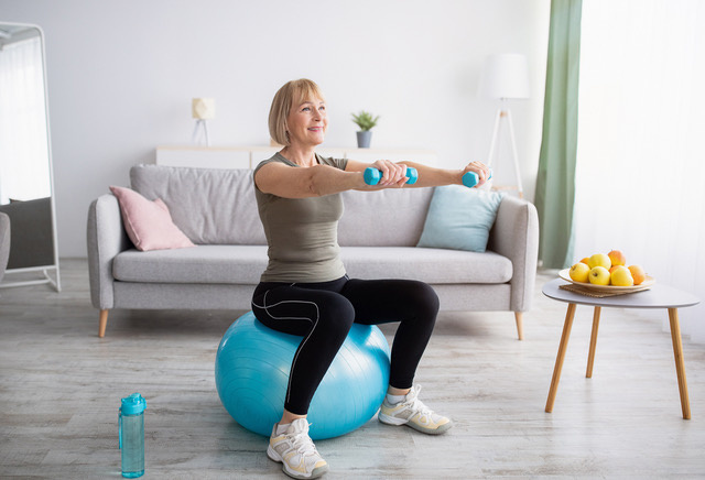 Woman is exercising in her living room with dumbbells and stability ball. 